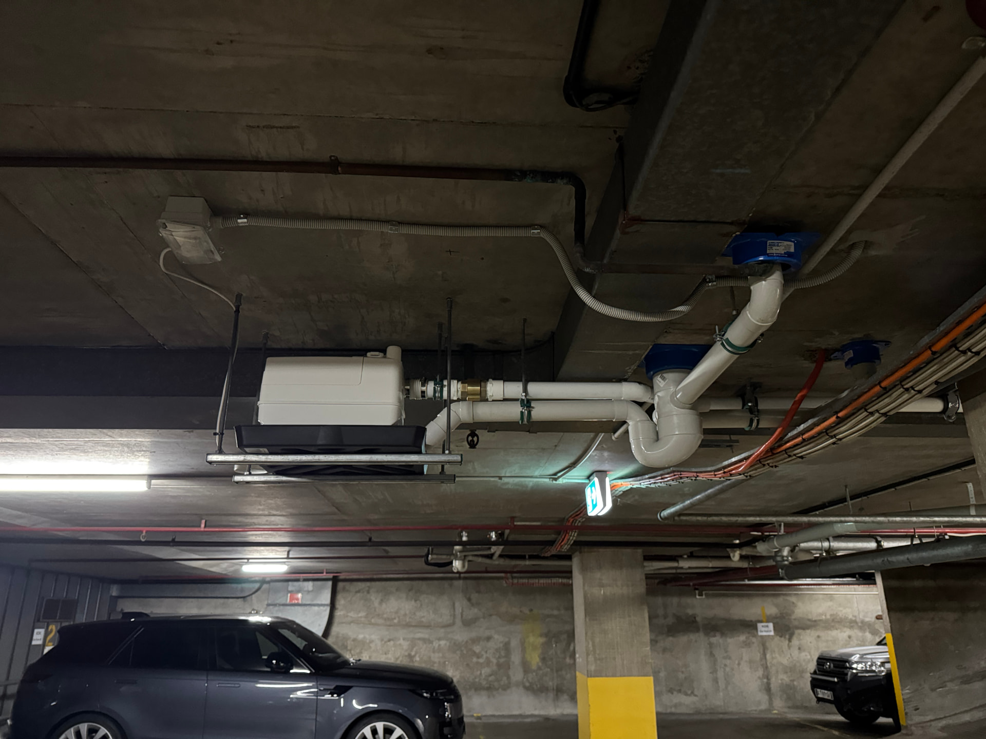 Macerator pump-out drainage unit strut-mounted to a basement car park soffit with discharge pipework and drip tray