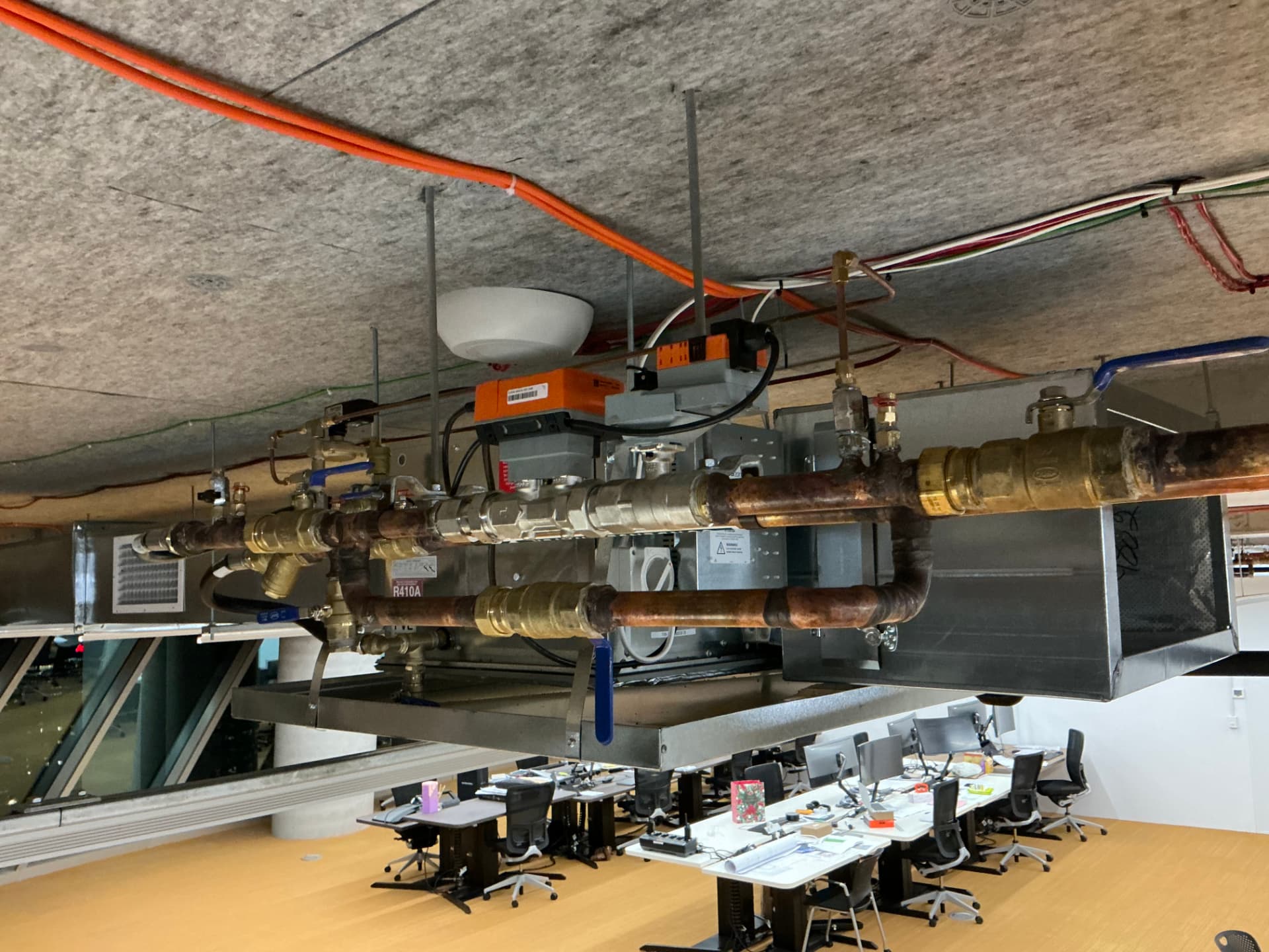 Copper chilled water pipework and brass valve manifold with motorised actuators connected to a ceiling-mounted PAC unit above a commercial office floor
